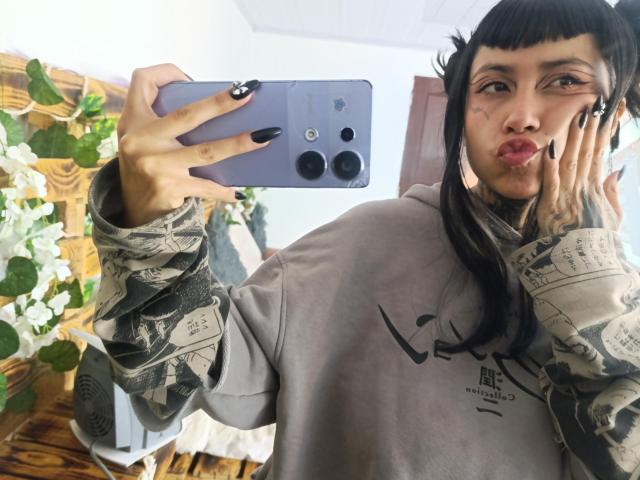 NyxVega taking a selfie with a purple phone, pouting lips, black nails, tattoos, long black hair, gray camouflage hoodie, holding phone in right hand, left hand on cheek.