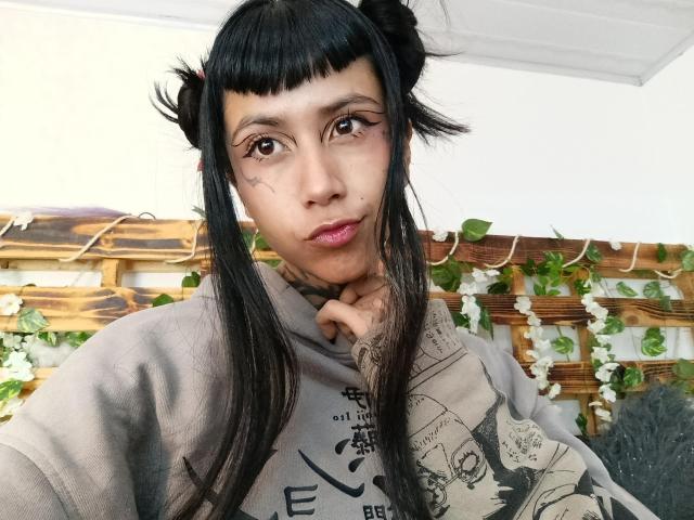 NyxVega with black double buns, straight black hair, brown eyes, tattooed neck, pink lipstick, grey graphic tee, resting chin on hand, looking at camera, cute expression.