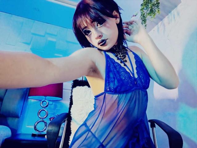 ViolettClay with short brown hair, blue lace babydoll, black choker, and dark lipstick, extending arm for selfie, showing armpit, chair, and blue lamp in background.