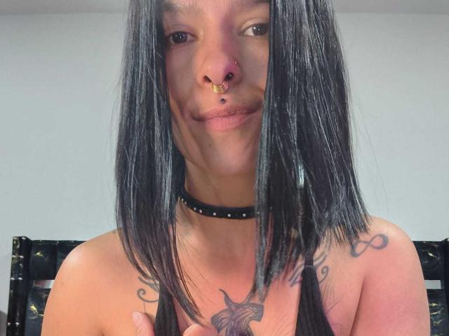 AmelieBlake with black straight hair, nose ring, lip piercing, black choker, tattooed shoulders, and visible chest. She's smiling, holding her tits, in a selfie.