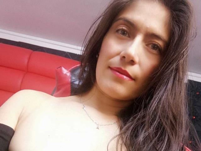 Sara Graham, mit dunkelbraunen Haaren, rotem Lippenstift und einer silbernen Halskette, hat freie Schultern und ein leichtes Lächeln und blickt in die Kamera. Ihre Brüste sind teilweise sichtbar..