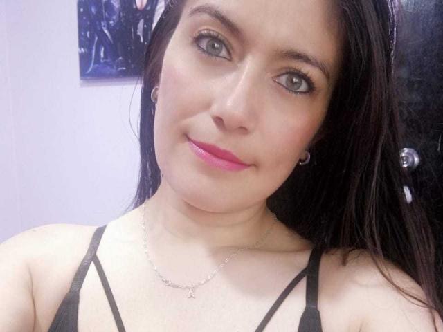 Sara Graham ha un seno di media grandezza, rossetto rosa, occhi verdi, capelli neri e lisci, indossa un top nero con spalline sottili, una collana d'argento e orecchini a cerchio, e sorride dolcemente alla fotocamera..
