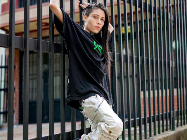 MorganEvans leans against black metal bars, wearing a loose black shirt, white pants, and green graphic, with long dark hair, looking slightly annoyed, in a street setting.