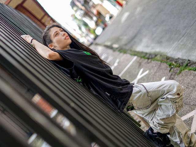 MorganEvans leans against a metal fence, wearing a black tee and white baggy pants, with long brown hair and a green necklace, looking up with a slightly open mouth.