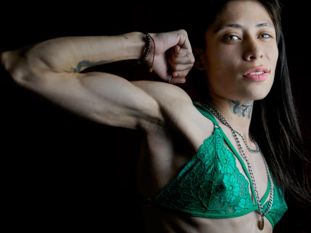 MorganEvans with muscular arms, green lace bra, tattoos, long black hair, chain necklace, and pink lipstick, flexing right arm against dark background. Boobs visible through lace.