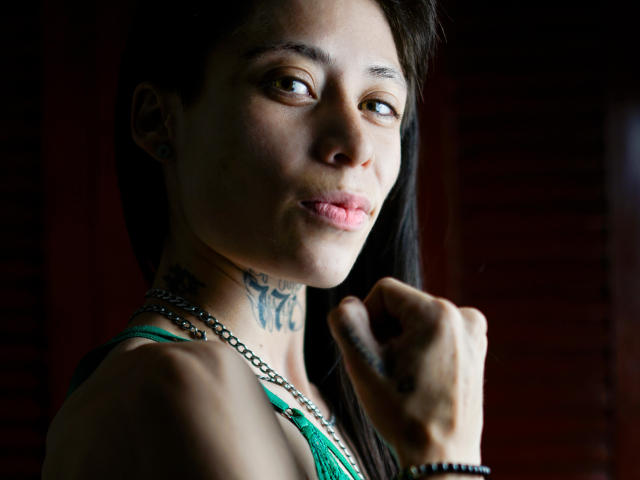 MorganEvans with pouty lips, green tank top, tattoos on neck, dark hair, brown eyes, holding fist, silver chain, black wristband, light skin, slight smile, dark background.