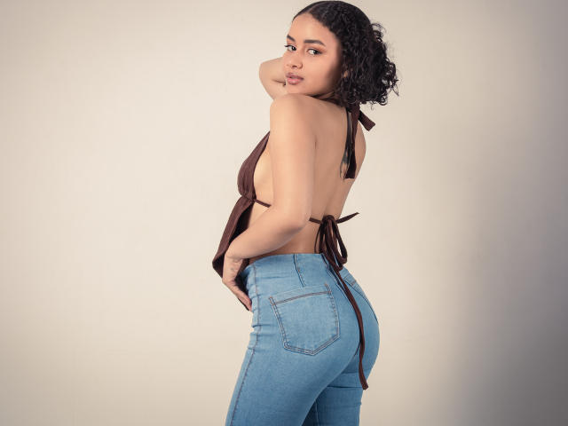 CurlyLorene with curly black hair, brown halter top revealing her back and sideboob, wearing blue jeans, looking over her shoulder, hand on her butt.