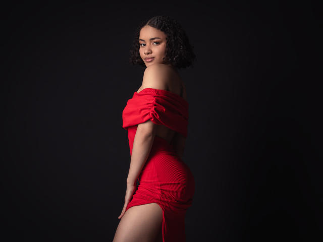 CurlyLorene with short curly black hair, wearing a red off-shoulder dress, showing her ass and a thigh slit, standing against a dark background, looking over her shoulder.