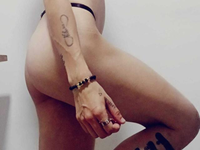 MorganEvans's bare butt, wearing black thong, showing tattoos on arm and thigh, bracelet on wrist, and rings on fingers, standing with one leg raised, in a photo.