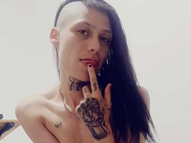 MorganEvans with shaved side, long black hair, tattoos on neck and hand, finger to lips, necklace, topless, pale skin, looking at camera, slight smile.