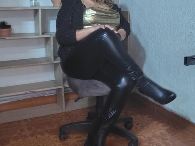 MatureYayita sitting on a chair with legs crossed, wearing black shiny leather boots, black pants, gold top, and black jacket, hands on lap, in a room.