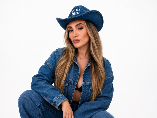 SashaEliteX, with long blonde hair, wearing a blue denim hat and jacket, slightly open to show her tits, squatting against a white background.