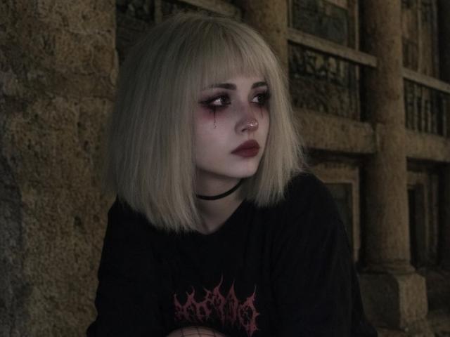 EmelyDilsaver has pale skin, a blonde bob, bloody black eye, dark lipstick, black choker, and a black shirt with a pink graphic. She's leaning against a stone wall.