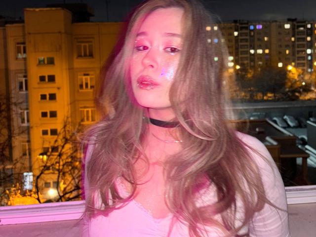 Siobhan Sunderland tem maquiagem rosa brilhante, cabelos castanhos longos e ondulados, um colar preto e uma blusa rosa, com um brilho rosado no rosto e no peito. Fundo da cidade à noite..