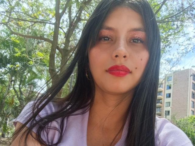 MadissonFontaine with long black hair, red lipstick, and shiny pink eyeshadow, wearing a purple shirt, standing outdoors with a tree and building in the background. Her face has a small beauty mark.