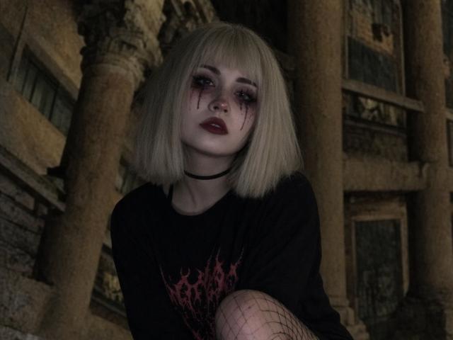 EmelyDilsaver with pale skin, bloody dark eye makeup, blonde bob, black choker, black t-shirt, fishnet on knee, standing in dark, gothic-looking building with columns.