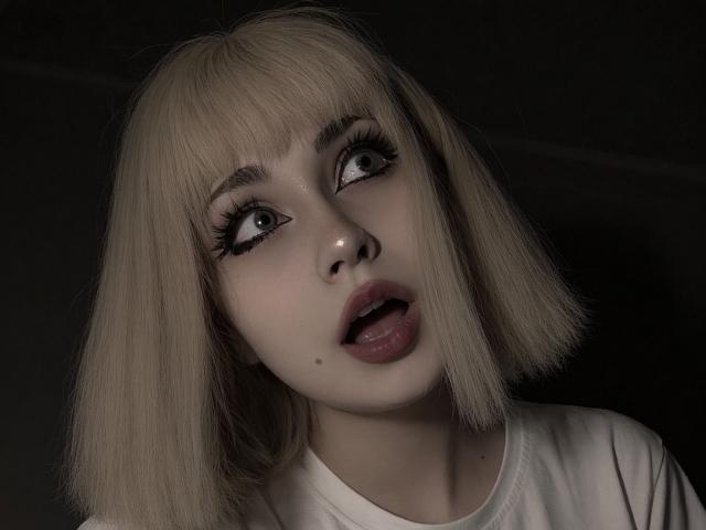 EmelyDilsaver has blonde bob, large green eyes, and thick black eyeliner. She's sticking her tongue out with her mouth slightly open, wearing a white shirt. Dark background.