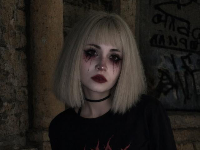 EmelyDilsaver with blonde bob, bloody black-eye, dark lipstick, nose ring, black choker, and black shirt with red graphic, looking serious in dim lighting.