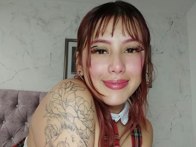 MaddyMills has pink eyeshadow, red lipstick, and a plaid dress. She's smiling, showing her puffy ass-tattooed shoulder with lilies. Her red hair has bangs.