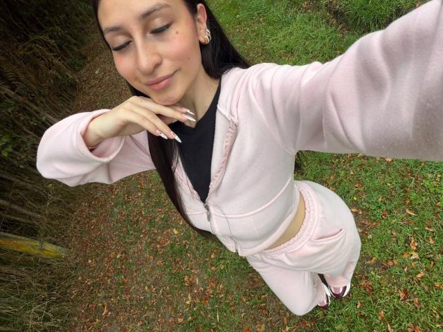 MaddyCarina with long dark hair, wearing a pink zip-up hoodie and matching pants, smiling, eyes closed, holding her chin, standing on green grass, showing her pink underwear.