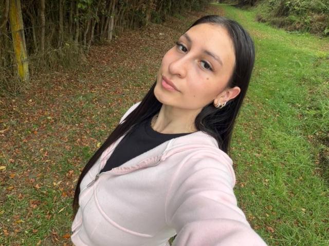 MaddyCarina with long black hair, wearing a pink zip-up hoodie and black top, taking a selfie on a grassy path, showing her small tits and a cute face.