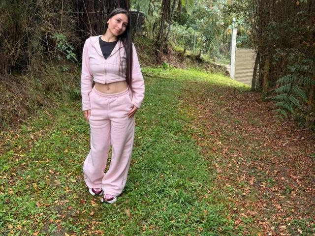 MaddyCarina stands in a forest, wearing pink pajama pants and a matching jacket, black top, hands in pockets, long black hair, smiling, green grass, brown leaves.