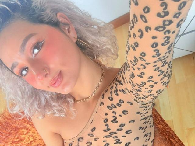 ElenaSaenz with curly white-blonde hair, wearing a leopard-print bodysuit, has pink blush, a nose ring, and a gold chain, smiling at the camera. Her arm is raised.