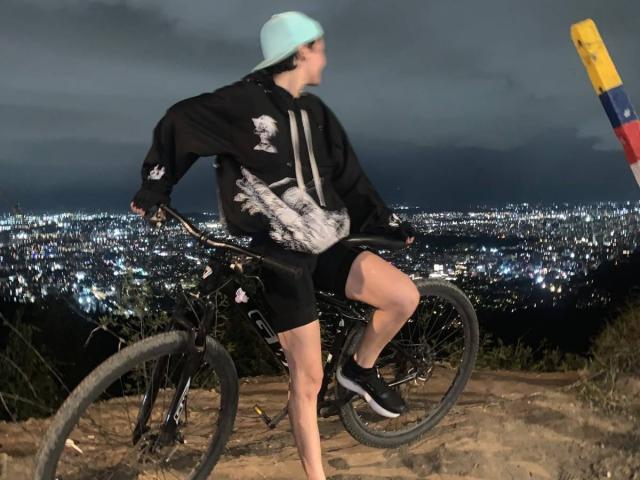 LissyLuxe with a blue cap, black hoodie, and shorts, riding a bike on a dirt trail at night, city lights in the background, wearing black shoes, and white socks.