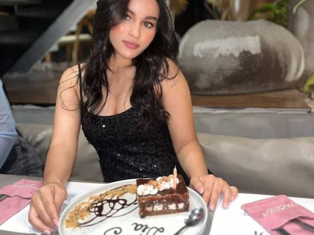 MiiaPalmer, avec ses longs cheveux foncés, portant une robe noire ornée de sequins, est assise à une table devant un gâteau au chocolat surmonté d'une bougie, et tient une fourchette..