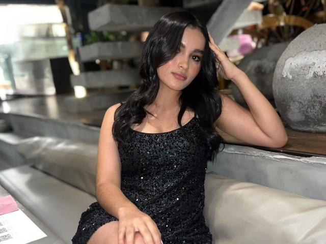 MiiaPalmer with long black hair, wearing a sparkly black dress, sitting on a white couch, hand in hair, looking at camera, medium shot, indoors.