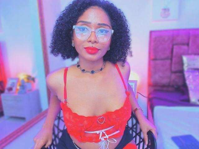 MeganWilliams with curly black hair, red lace top, heart button, and red lipstick, wearing clear glasses and a necklace, sitting on a chair in a brightly lit room.