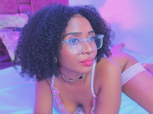 MeganWilliams with curly black hair, wearing glasses, a pink lace bra, and a white lace thong, lying on a bed with blue and pink lighting. Boobs, ass, and a choker.