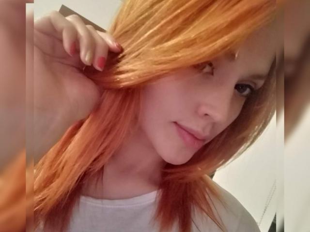 LinaStars with orange hair, slightly smiling, holding her hair with one hand, wearing a white shirt, showing pink-nailed fingers, and a cute, relaxed face.