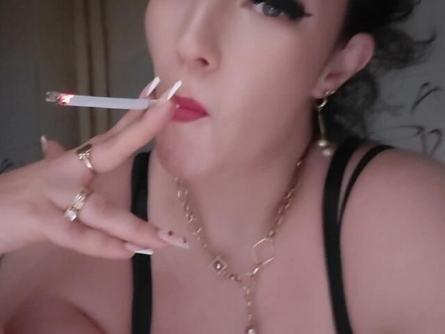 VanessaSquirtt with long nails, red lipstick, and gold jewelry, holding a lit cigarette near her mouth, wearing a black top, showing her tits.