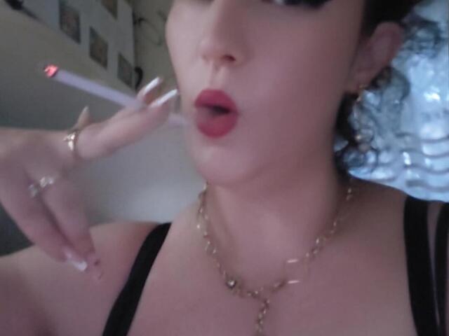 VanessaSquirtt with red lips, holding a lit cigarette near her mouth, wearing a black top and gold necklace, showing her ringed fingers and a slight pout.