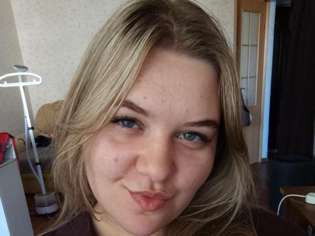 DelmaMoen has blonde hair, blue eyes, and is making a pouty face with slightly puckered lips. She has a few acne spots on her face and is wearing a brown shirt.
