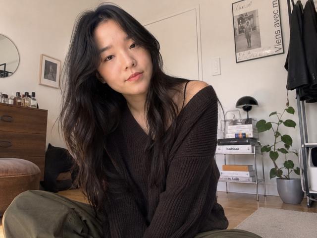 SamanthaTango with long black hair, wearing an off-shoulder brown knit sweater, green pants, sitting on floor, looking at camera, soft smile, light skin, cute.