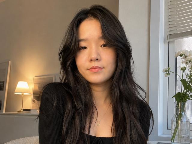 SamanthaTango with long, wavy black hair, light skin, and a slight smile, wearing a black ribbed shirt, looking at the camera, in a softly lit room.