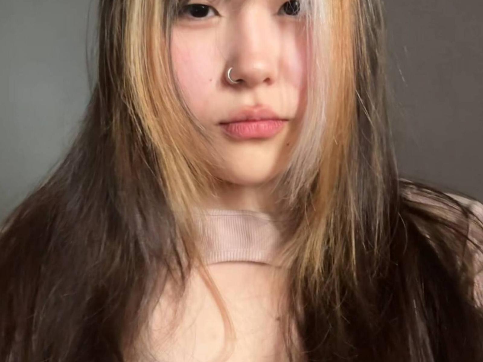 JungMcgranor tiene el cabello largo y castaño oscuro con reflejos rubios, un piercing en la nariz y labios rosados, y lleva un collar beige. Su rostro está centrado y mira ligeramente hacia abajo..
