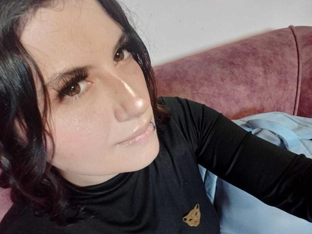 MaryCruzz with black curly hair, light skin, and subtle makeup, wearing a black shirt, showing a small piercing on her lower lip, sitting on a pink sofa.