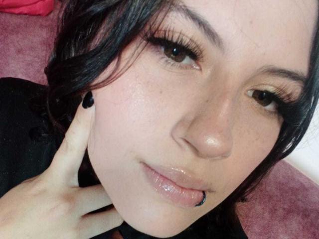 MaryCruzz has dark wavy hair, long eyelashes, and glossy pink lips. She's sucking a black-chipped nipple with her finger touching her cheek. Her eyes are slightly up.
