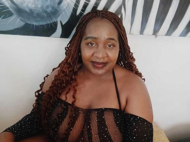 SexcCoco, a curvy Black woman with long red braids, wearing a sheer black top, has large tits and a warm smile, sitting on a white couch.