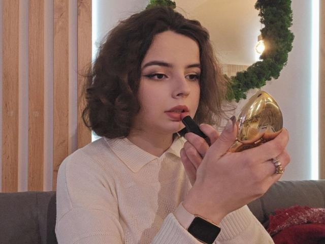 MiamMooon with curly brown hair, applying dark lipstick to her plump, pink lips with a gold, oval lipstick holder in her hand, wearing a white, ribbed sweater.