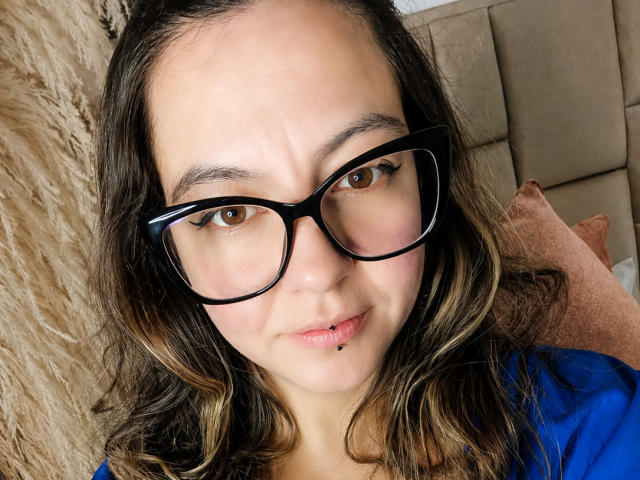 ArianaBlonde has black glasses, brown eyes, wavy dark blonde hair, a lip piercing, and a blue shirt. Her face is slightly squished with a neutral expression.