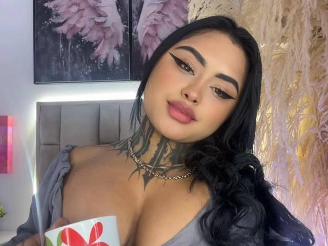 LairaHearvy with black hair, thick eyebrows, and pink lipstick, showing off her big tits in a grey off-shoulder top, holding a red heart card.