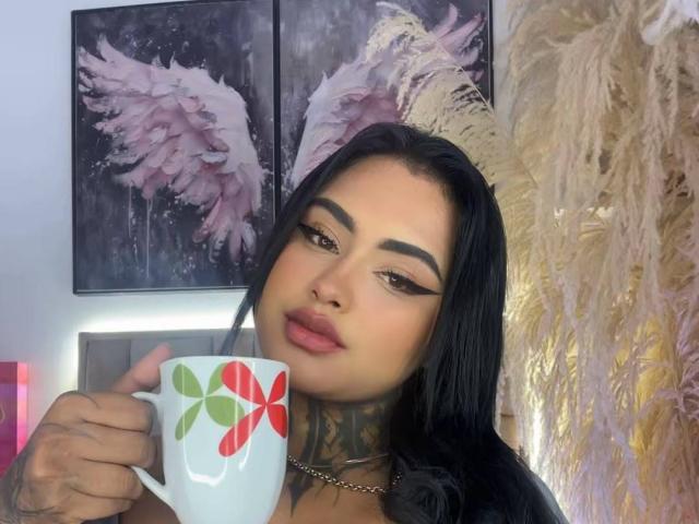 LairaHearvy with black hair, bold eyebrows, and red lipstick holding a white mug with red and green flower patterns. She has tattoos on her neck and chest.
