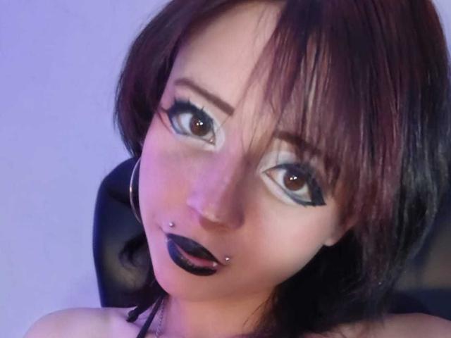 ViolettClay with dark brown eyes, black eyeliner, and black lipstick, has a nose piercing and lip piercing, with short brown hair, wearing black hoop earrings.