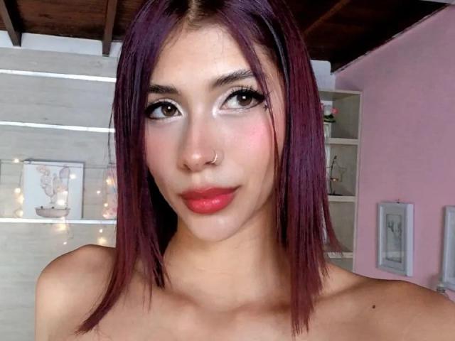 DanikaSerov with purple hair, nose ring, and pink lips, showing off her tits, looking at the camera with a slight smile, in a well-lit room.