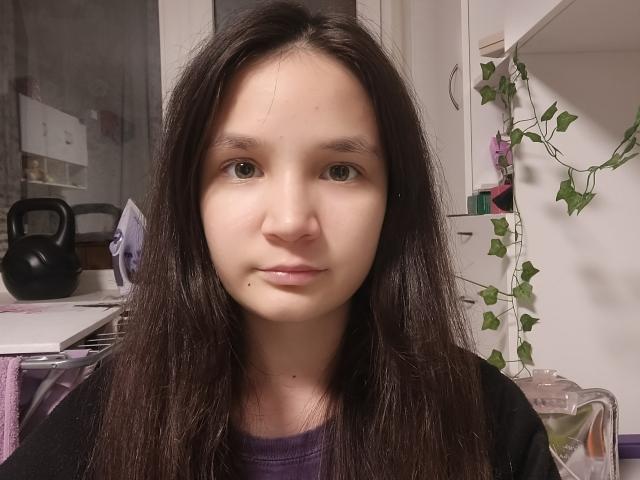 Odelia Holen hat langes, dunkelbraunes Haar, helle Haut, kleine Brüste und einen Muttermal unter ihrer rechten Lippe. Sie trägt ein lila Hemd und ein schwarzes Oberteil..