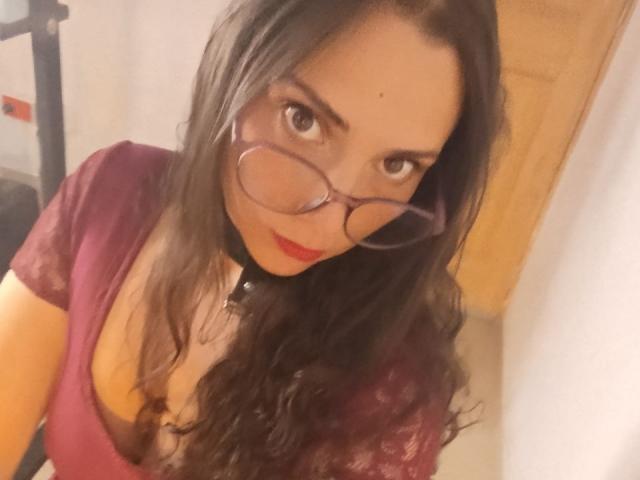 VelhMurphi, con lunghi capelli scuri e ondulati, indossa occhiali viola, rossetto rosso e una camicetta di pizzo viola scuro, che lascia intravedere un po' il seno. Sta guardando verso l'alto, verso la fotocamera..
